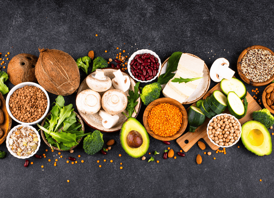 Assorted fresh vegetables, legumes, seeds, tofu, and avocado arranged on a dark surface, viewed from above.
