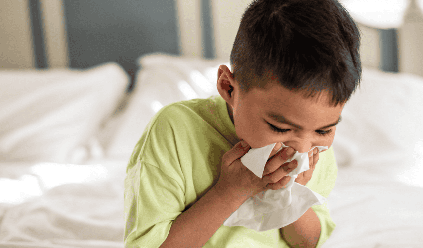 A young boy in a light green shirt sits on a bed and blows his nose into a tissue.