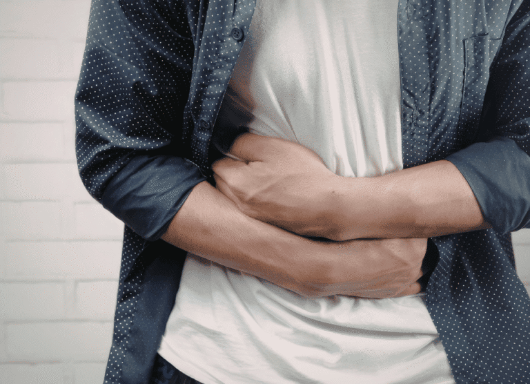 A person holding their stomach with both hands, wearing a white shirt and blue polka dot overshirt, standing against a white wall.