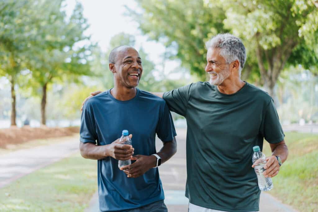 Two men in athletic wear walk outdoors on a path, smiling and holding water bottles, with trees and grass in the background.