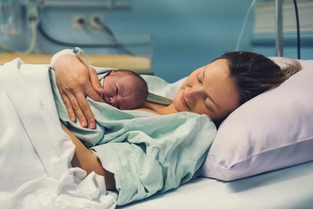 A woman lies in a hospital bed, holding a newborn baby on her chest, both covered with a light blanket.