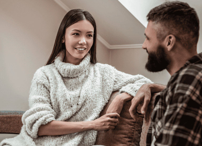 A woman in a light sweater sits on a couch, talking to a man in a plaid shirt who is sitting nearby.