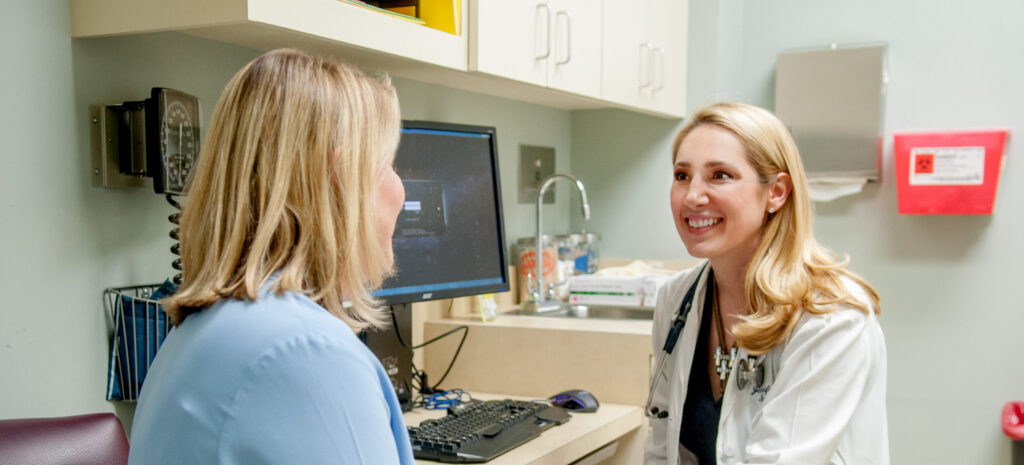 A doctor in a white coat talks with a seated patient in a medical office with a computer and medical equipment visible.