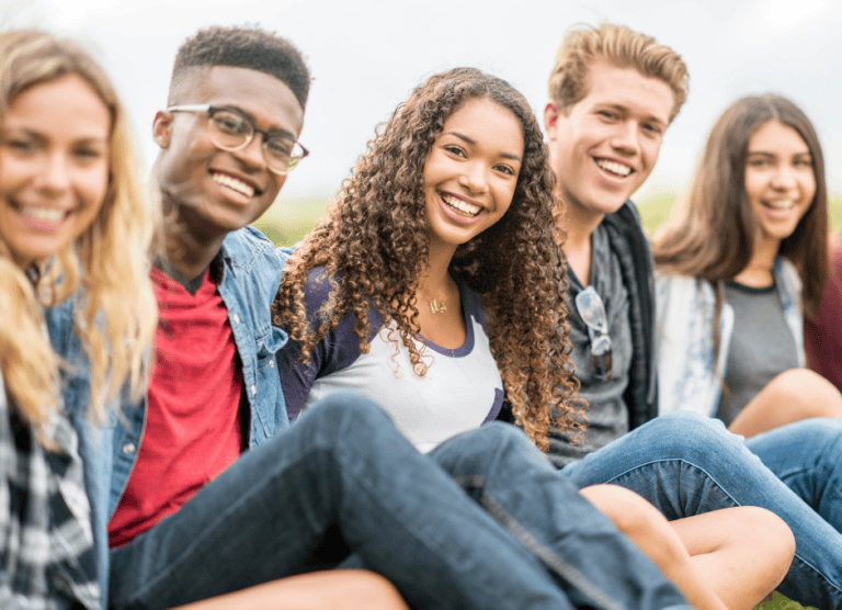 Five young adults sit outdoors in a line, smiling at the camera on a bright day.