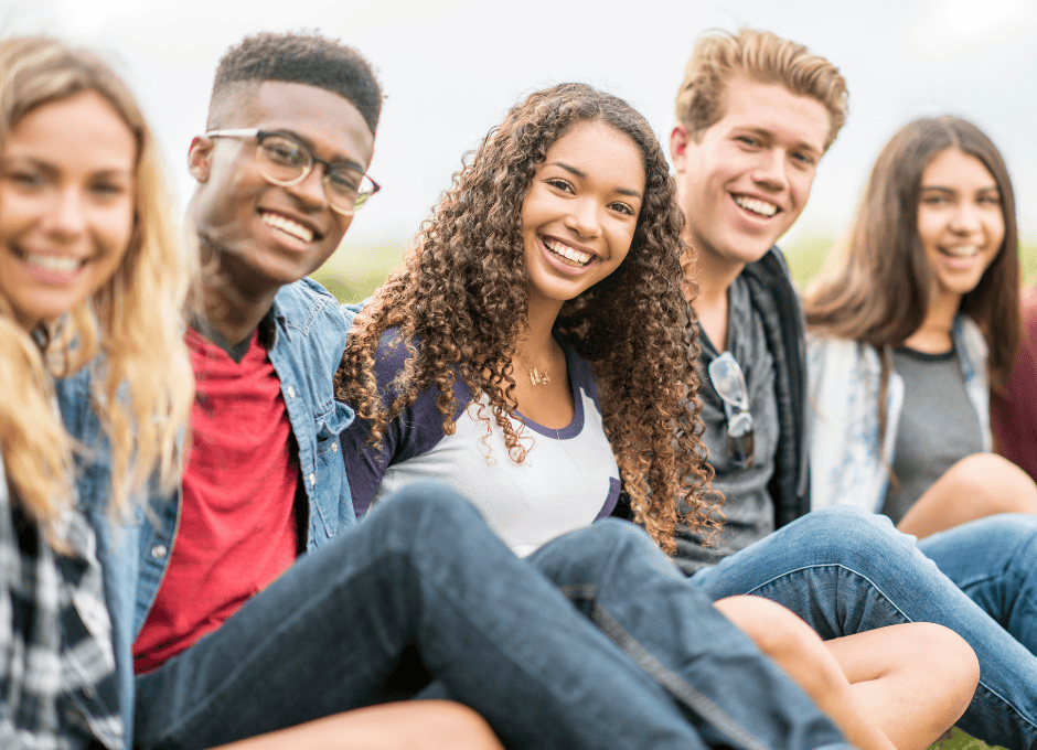 Five young adults sit outdoors in a line, smiling at the camera on a bright day.