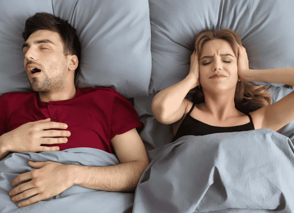 A man is sleeping and snoring while lying on his back. A woman next to him covers her ears and appears frustrated.