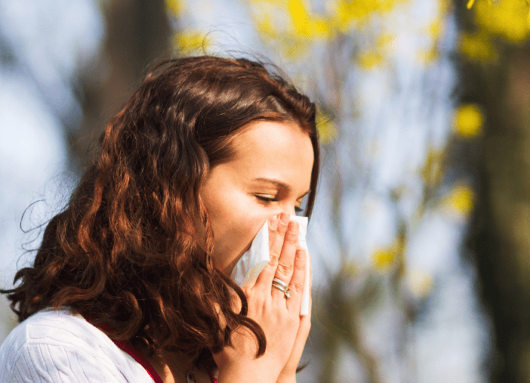 A woman with brown hair holds a tissue to her nose, appearing to sneeze, with trees and yellow flowers in the blurred background.