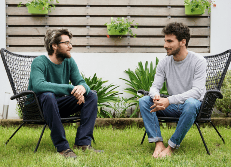 Two men sit on black chairs in a garden, facing each other and talking, with plants and a wooden fence in the background.