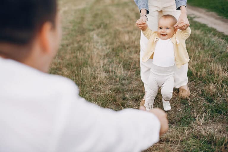 A baby walks on grass with help from an adult holding their hands; another adult reaches out in the foreground.