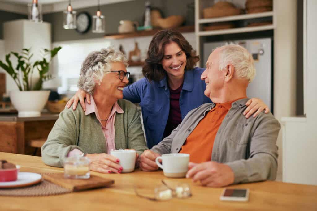 An older couple sits at a kitchen table with coffee while a younger woman stands behind them, smiling and talking.